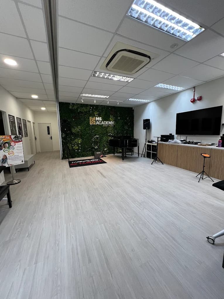 Open corridor and common space at MS Academy with a black grand piano, a wall-mounted TV, and the green MS Academy logo wall in the background