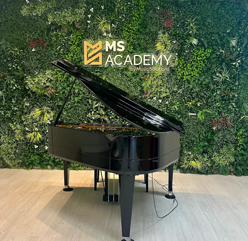Close-up of a black grand piano with the lid open, standing against the green MS Academy logo wall